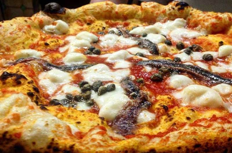 Una pizza con mozzarella a pezzi irregolari, filetti di acciuga e capperi su fondo di pomodoro.