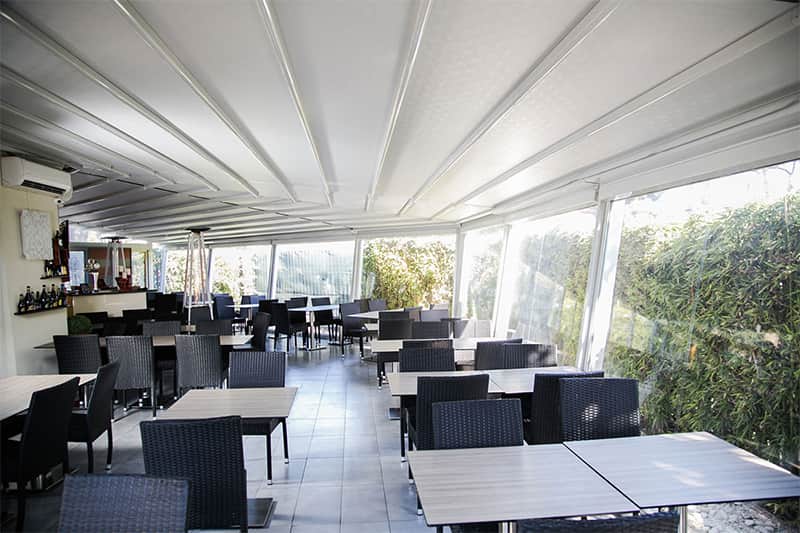 Sala interna con tavoli in legno e sedie intrecciate scure, pareti in vetro affacciate sul verde e soffitto bianco a pieghe.