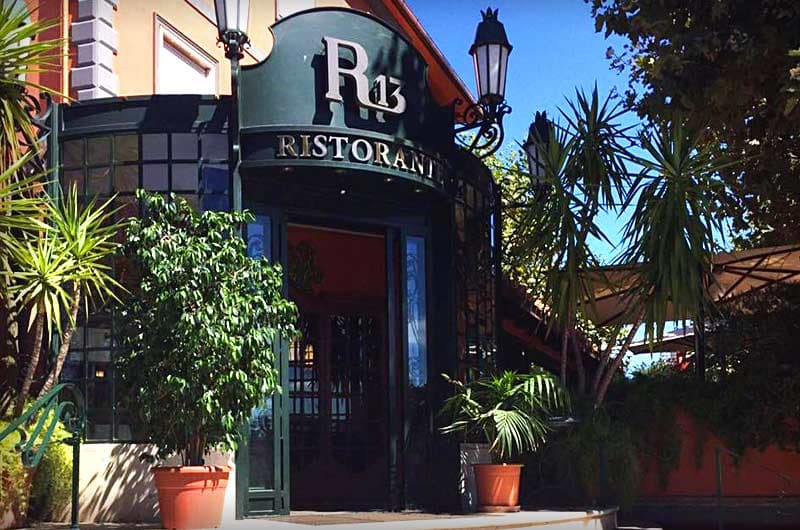 Facciata curva in vetro e metallo verde scuro con insegna dorata «R13 Ristorante» sopra la porta d'ingresso aperta.