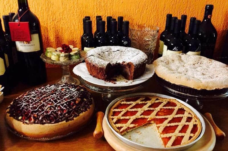 Crostate e torte disposte su un tavolo, con bottiglie di vino rosso allineate su sfondo arancione.