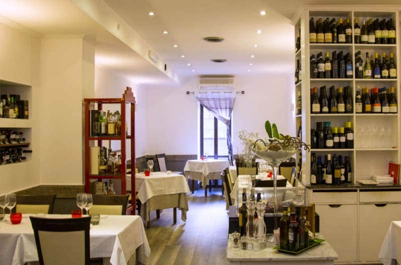 Sala con tavoli bianchi apparecchiati e scaffali a muro pieni di bottiglie di vino.