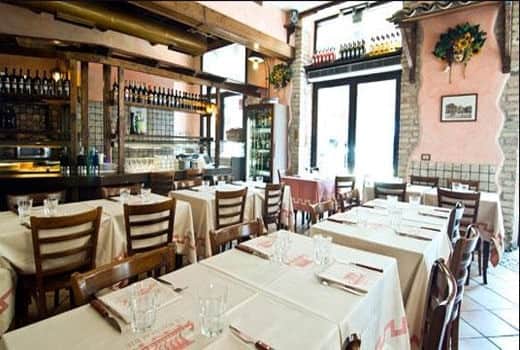 Sala con tavoli apparecchiati di bianco, sedie in legno e un bancone con bottiglie di vino sullo sfondo.