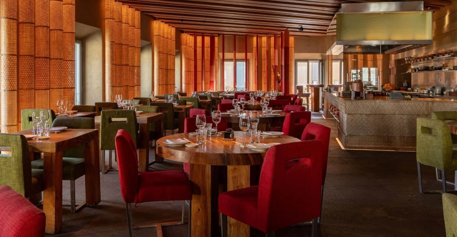Sala di ristorante con tavoli in legno, sedie rosse e verdi, pareti in canne intrecciate e cucina a vista sullo sfondo.