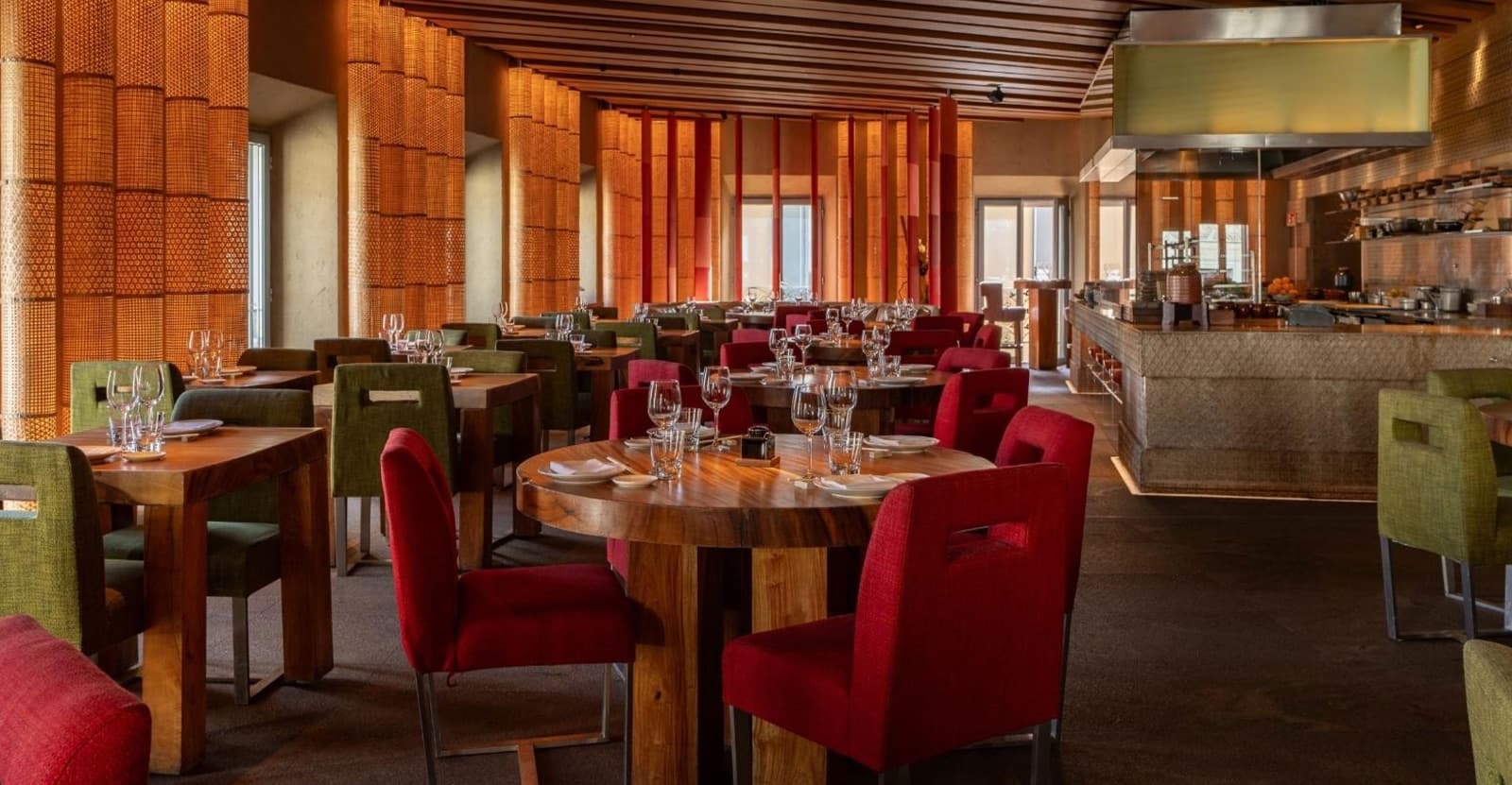 Sala di ristorante con tavoli in legno, sedie rosse e verdi, pareti in canne intrecciate e cucina a vista sullo sfondo.