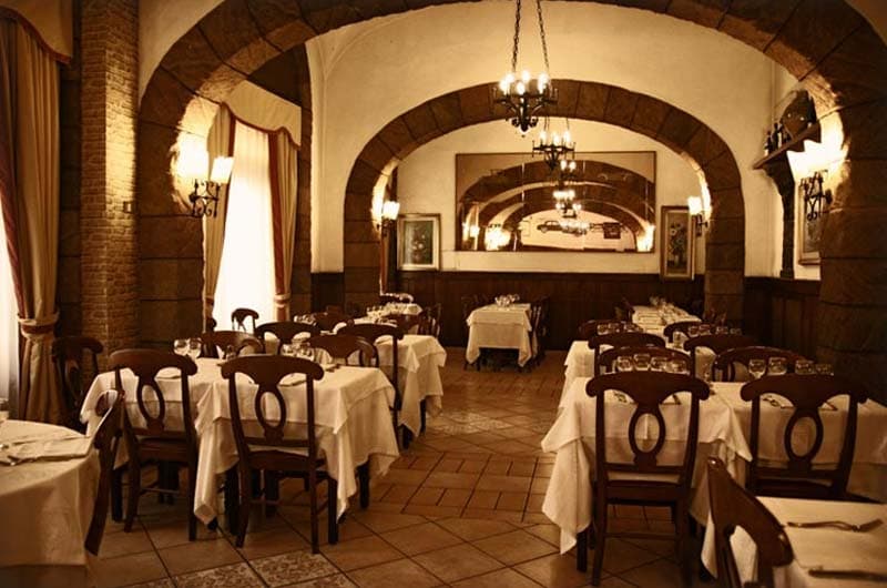 Sala con archi in mattoni, tavoli con tovaglie bianche e lampadario a bracci con candele.