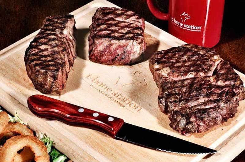 Tre bistecche grigliate su un tagliere di legno, con coltello a manico rosso, anelli di cipolla e una tazza rossa sullo sfondo.