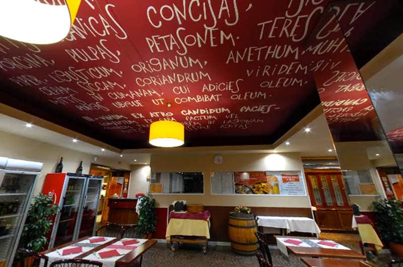 Soffitto rosso scuro con scritte bianche in latino su erbe e spezie, sopra una sala con tavoli apparecchiati.