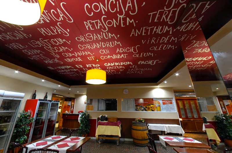 Soffitto rosso scuro con scritte bianche in latino su erbe e spezie, sopra una sala con tavoli apparecchiati.