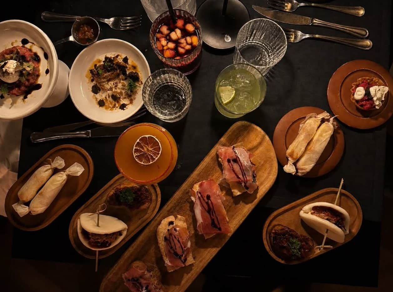 Piccoli piatti e cocktail visti dall'alto su un tavolo scuro, tra cui bruschette con prosciutto e panini al vapore.
