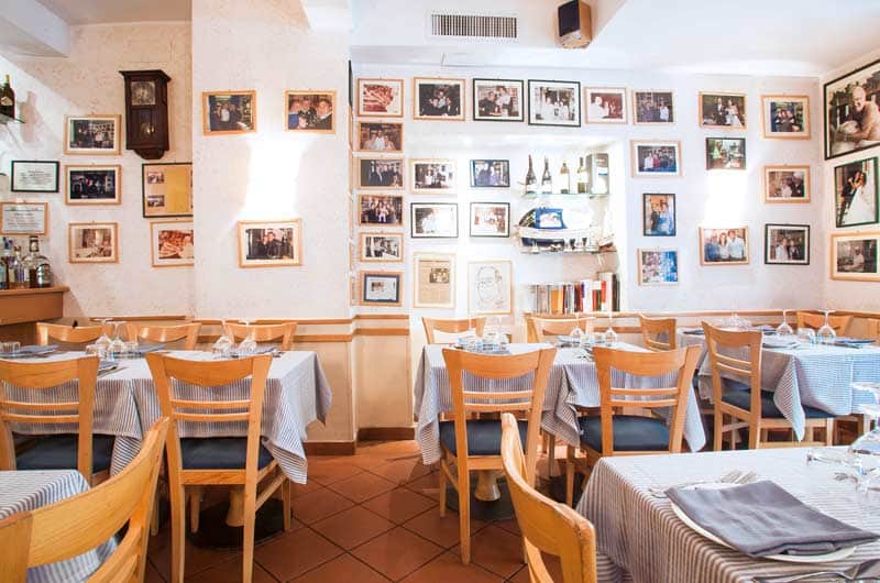 Sala di ristorante con pareti coperte di foto incorniciate, tavoli con tovaglie a righe blu e sedie in legno con seduta blu.