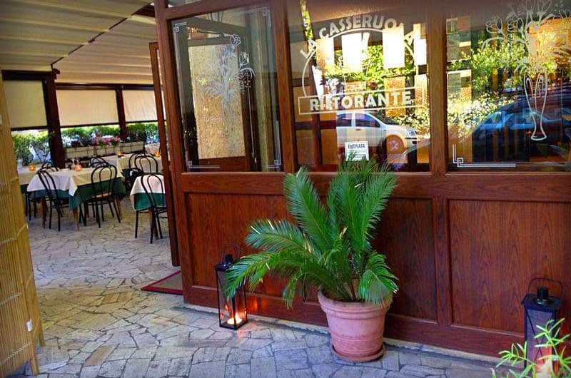 Ingresso di un ristorante con vetrata scritta, porta a vetri aperta e una pianta in vaso di terracotta davanti.