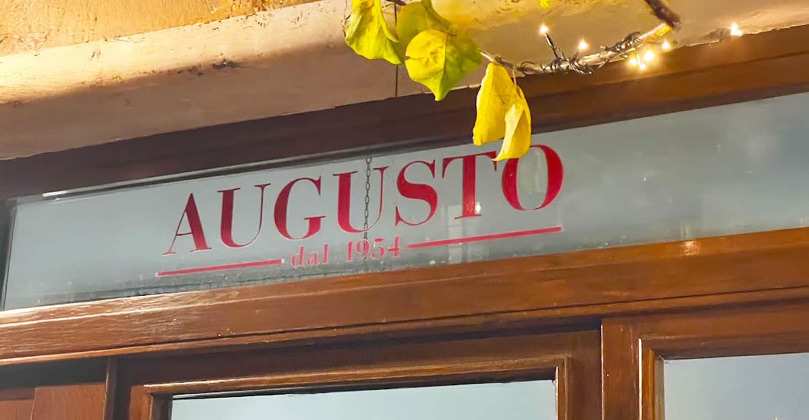 Insegna in vetro con la scritta Augusto dal 1954 in rosso, sopra una porta di legno con rami e foglie gialle appese.