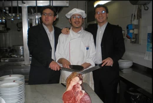 Cuoco in giacca e cappello bianco taglia un pezzo di carne cruda su un bancone, affiancato da due uomini in giacca e cravatta.