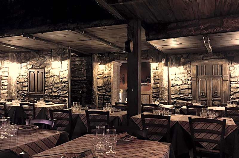 Sala con pareti in pietra, tavoli con tovaglie a quadri e bicchieri, illuminata da lampade a muro.