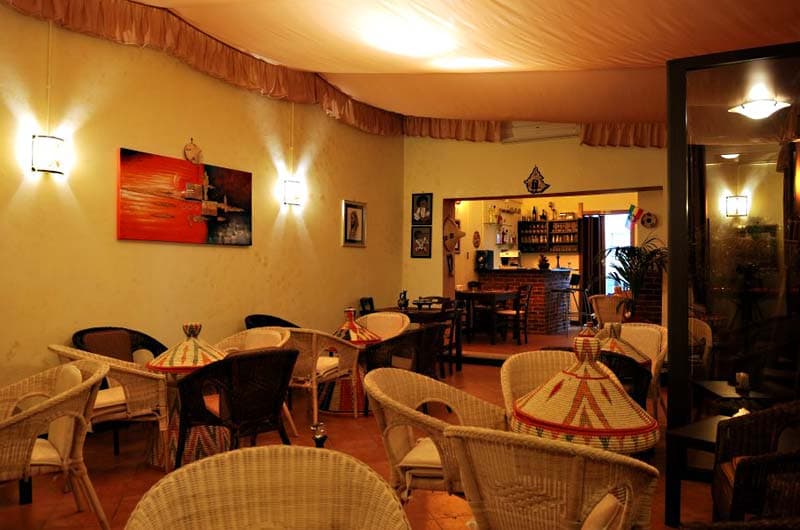 Sala con poltrone e sedie in vimini, tavoli con cesti conici decorati a righe rosse, verdi e gialle, luce calda da applique a muro.