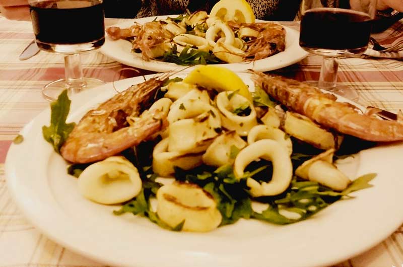 Piatto con gamberi e anelli di calamaro grigliati su rucola, con spicchio di limone, due bicchieri di vino rosso sullo sfondo.