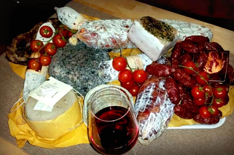 Tagliere con salumi, formaggi, pomodorini e un bicchiere di vino rosso su carta gialla.