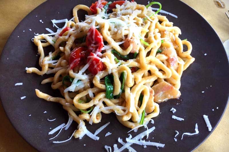 Piatto di pasta al nido con pomodorini, foglie verdi e formaggio grattugiato, su piatto scuro.