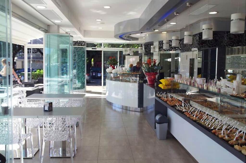 Sala con banco vetrina di dolci e gelati a destra, tavoli bianchi e sedie trasparenti a sinistra.