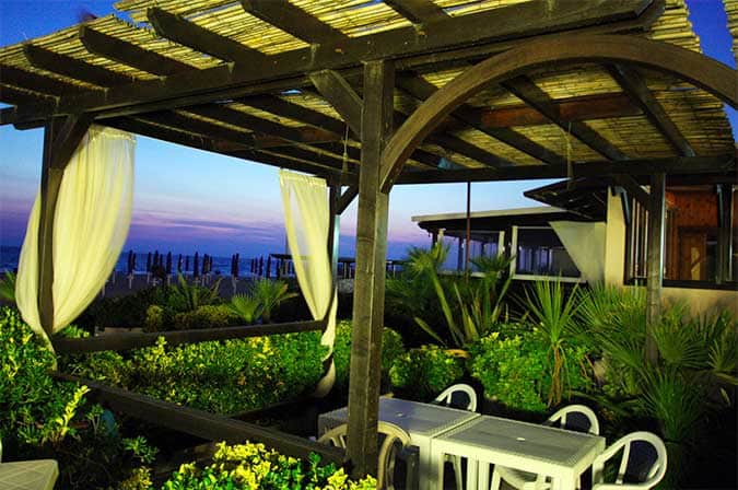 Pergolato in legno con tende bianche e tavolini apparecchiati, sullo sfondo la spiaggia al tramonto.