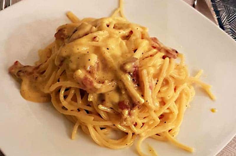 Spaghetti con salsa cremosa e pancetta, conditi con pepe nero, su piatto quadrato bianco.