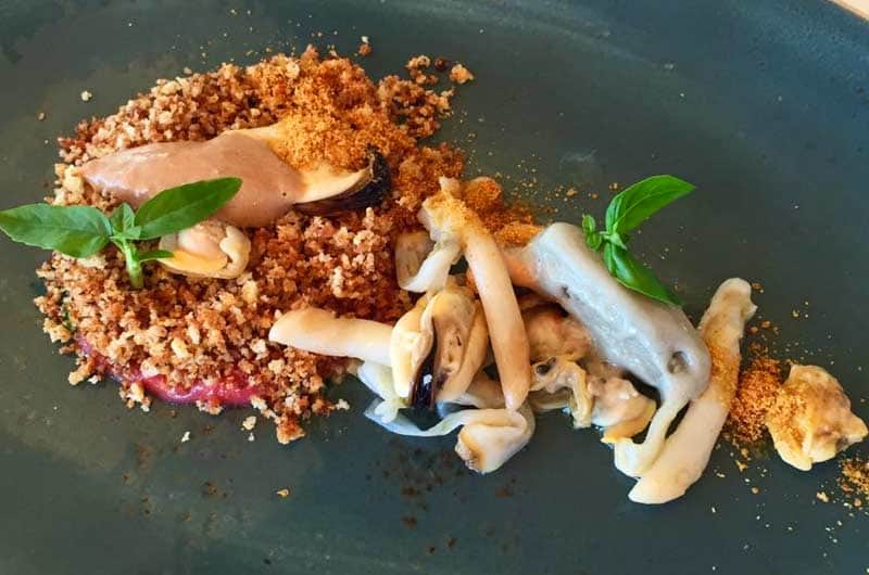 Piatto scuro con mollica sbriciolata, cozze, strisce di frutti di mare e una crema chiara, decorato con foglie di basilico.