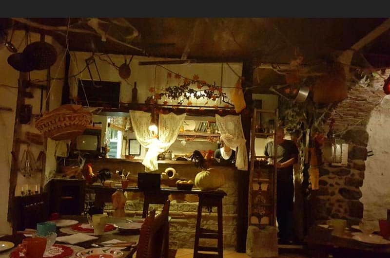 Sala rustica in penombra con travi in legno, tende bianche, zucche decorative e un bancone in pietra sullo sfondo.