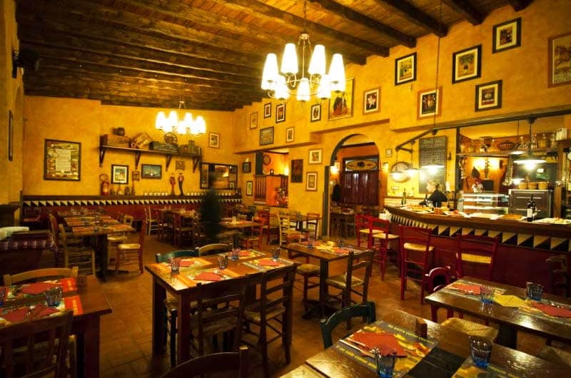Sala da ristorante con travi in legno a vista, tavoli quadrati apparecchiati e lampadario a bracci acceso al centro.