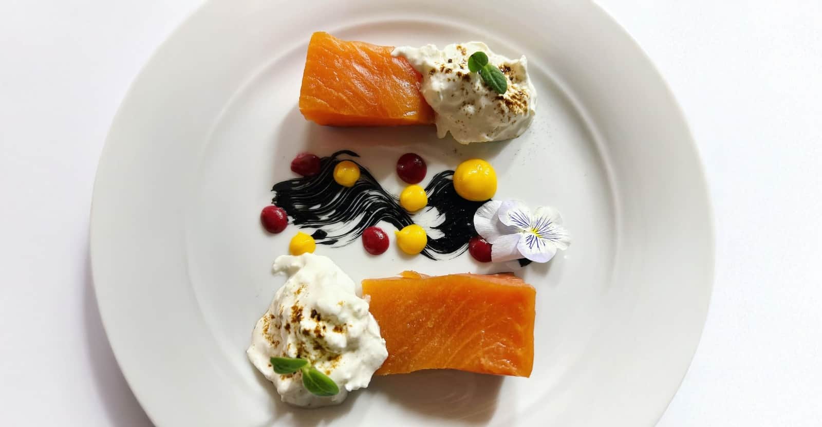 Due porzioni di salmone arancione con una quenelle di formaggio cremoso, decorate con pennellate nere, punti gialli e rossi e un fiore.