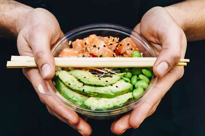 Due mani reggono una ciotola di poke con salmone a cubetti, avocado a fette ed edamame, con bacchette appoggiate sopra.