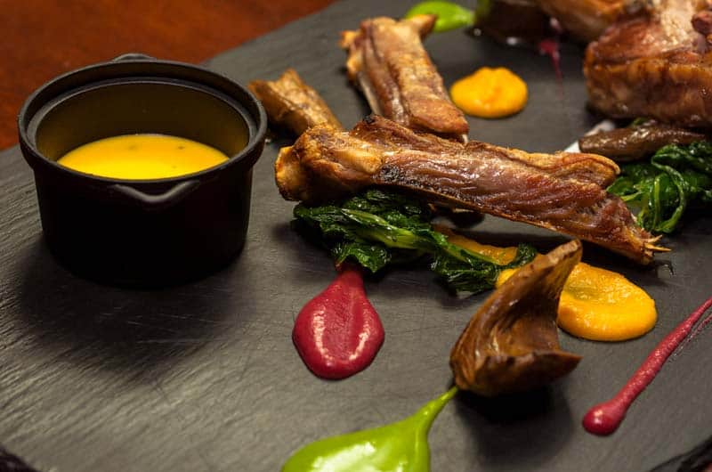 Costine di carne alla griglia su foglie di verdura saltata, con salse a specchio color rosso, giallo e verde su lastra scura.