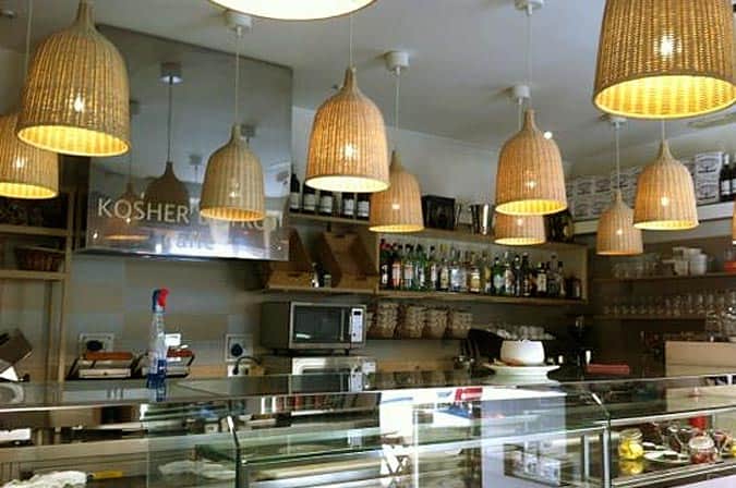 Lampade a cono in vimini pendono dal soffitto sopra un bancone con scaffali di bottiglie e uno specchio con la scritta KOSHER.