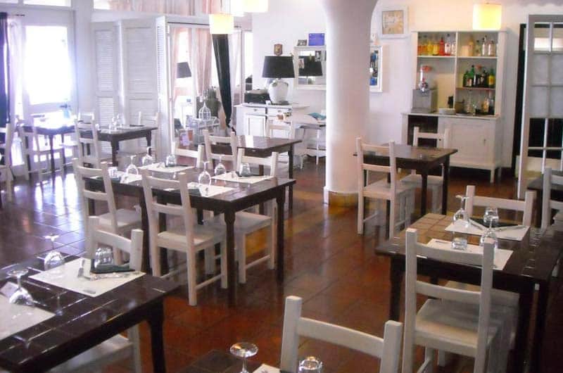 Sala con tavoli in legno scuro e sedie bianche apparecchiati con bicchieri da vino, sullo sfondo una credenza con bottiglie.