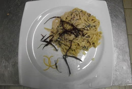 Piatto quadrato bianco con spaghetti conditi da striscioline scure e scorza di agrumi arricciata.