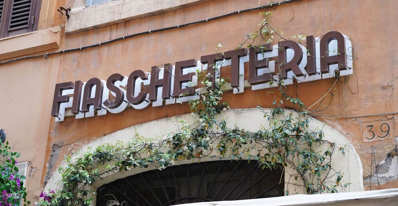 Insegna in rilievo con la scritta FIASCHETTERIA su una facciata color ocra, sopra un arco con rampicanti in fiore.