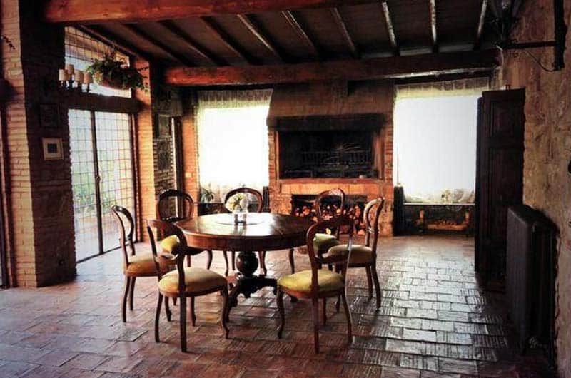 Sala da pranzo con travi in legno a vista, tavolo rotondo con sei sedie e un grande camino in mattoni.