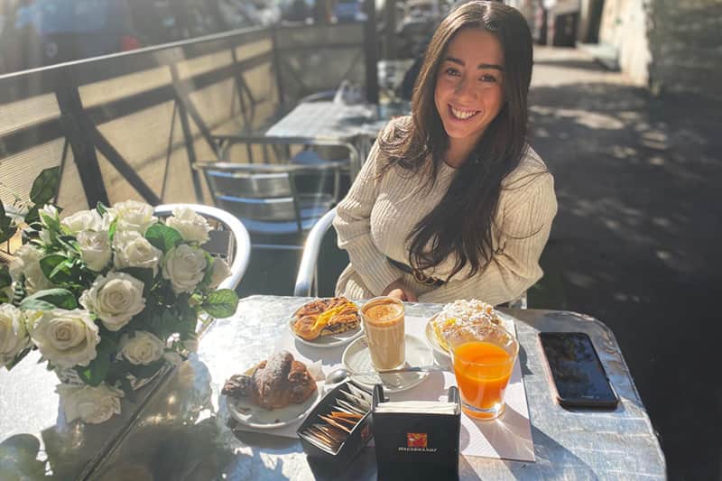Donna sorridente seduta a un tavolo con cappuccino, cornetto, un dolce con panna e un succo d'arancia, vaso di rose bianche a sinistra.