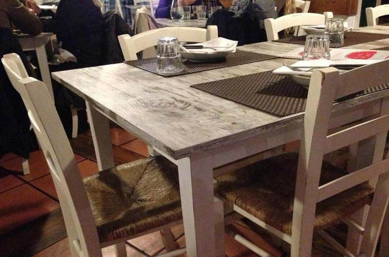 Tavolo in legno chiaro apparecchiato con tovagliette scure, bicchieri e tovaglioli.