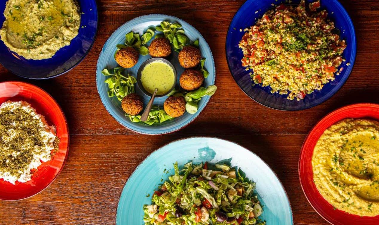 Sei piatti colorati su tavolo di legno scuro con falafel, hummus, tabulè e insalata mista.