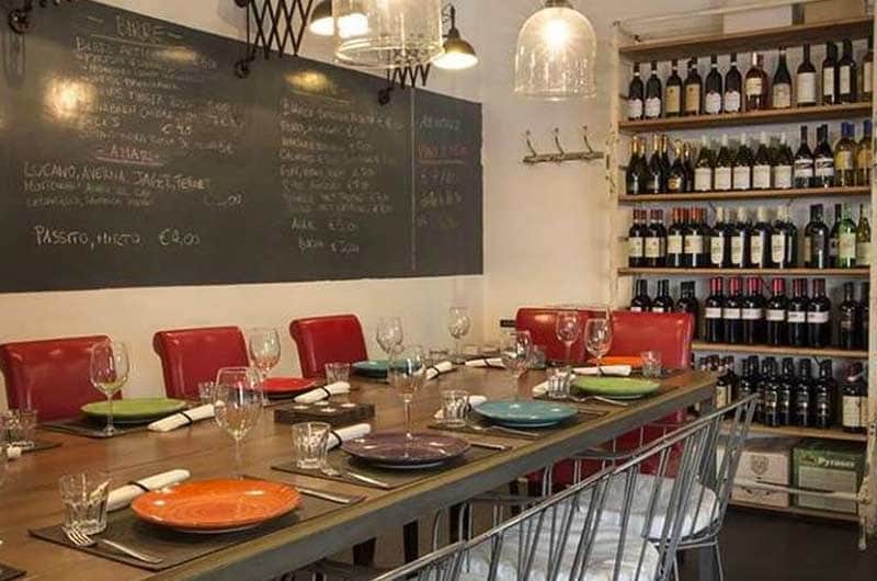 Tavolo lungo con piatti colorati e bicchieri da vino, lavagna con menu scritto a mano e scaffali di bottiglie sullo sfondo.