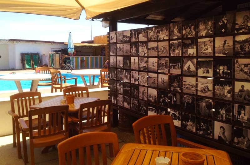Dehors con tavoli in legno, parete ricoperta di fotografie in bianco e nero e piscina sullo sfondo.
