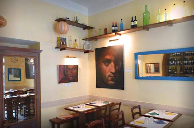 Sala con pareti gialle, mensole con bottiglie di vetro colorato e due grandi ritratti dipinti sopra i tavoli apparecchiati.