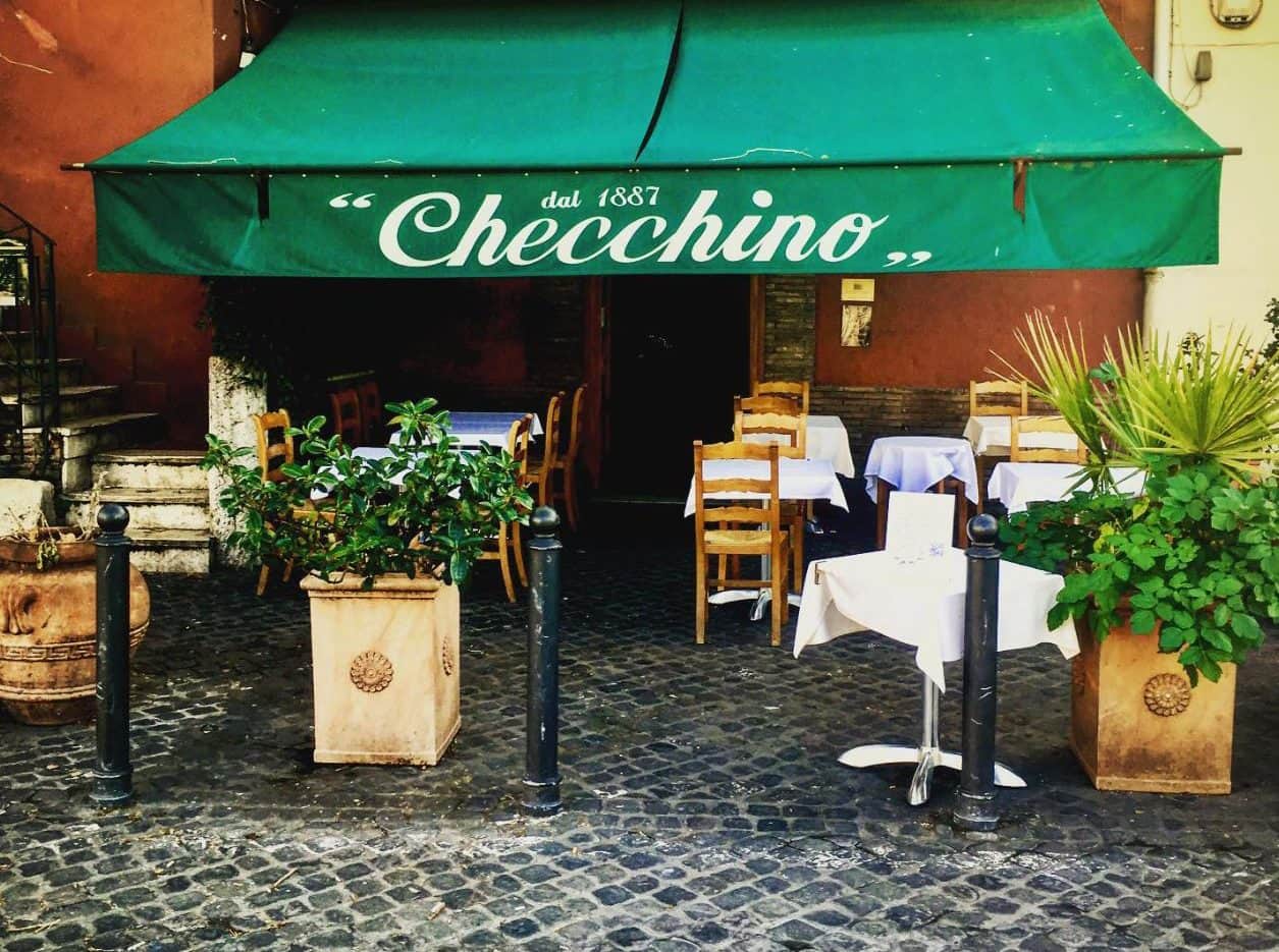 Dehors con tavoli bianchi e sedie di legno sotto un'insegna verde con la scritta Checchino dal 1887.