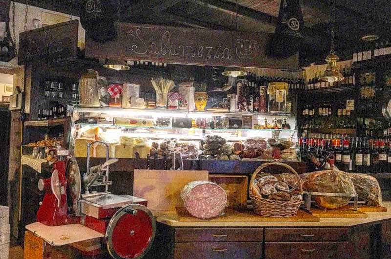 Bancone di salumeria con vetrina illuminata di formaggi e affettati, salame e prosciutto sul piano di legno.