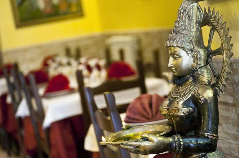 Statua dorata di divinità indiana con vassoio e lampada, in primo piano davanti a tavoli apparecchiati con tovaglie bianche.