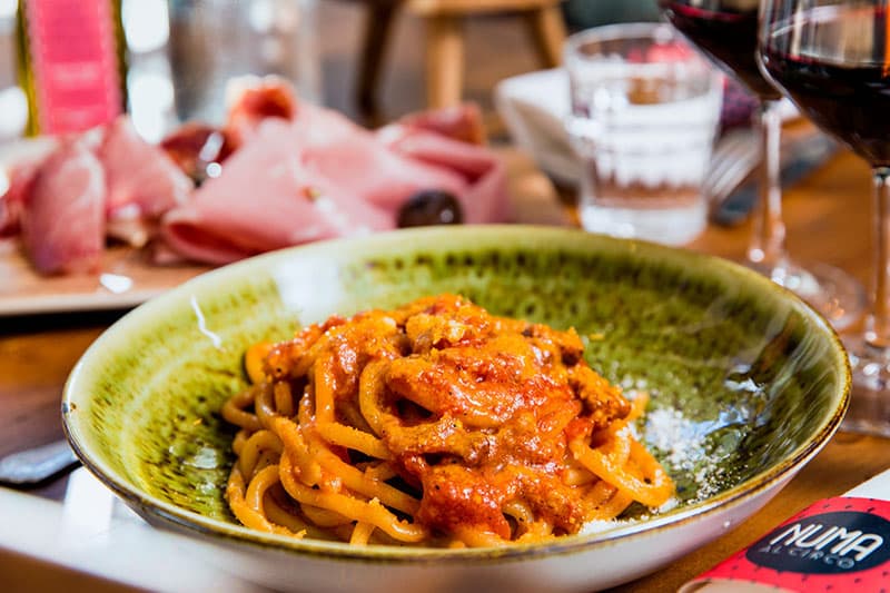 Ciotola verde con pasta lunga in sugo rosso, sullo sfondo salumi affettati e calici di vino rosso.