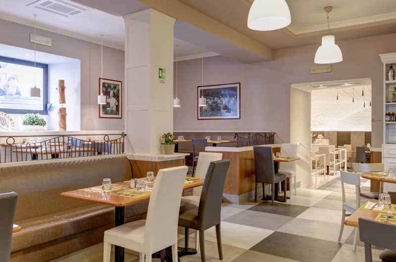 Sala da ristorante con colonna bianca centrale, panche imbottite, tavoli in legno e pavimento a scacchi grigio e bianco.