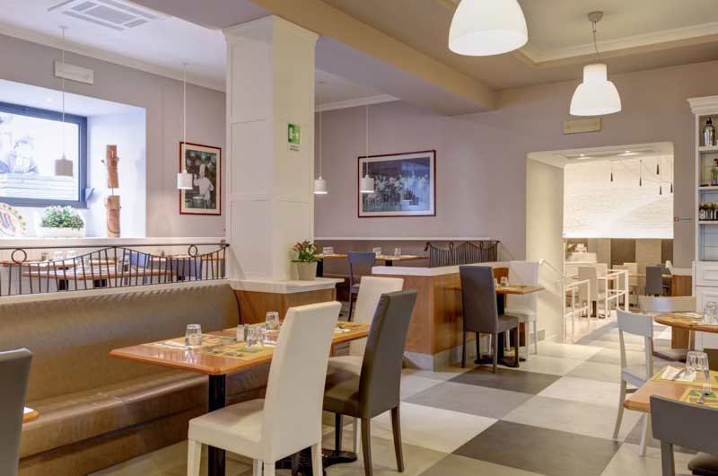 Sala da ristorante con colonna bianca centrale, panche imbottite, tavoli in legno e pavimento a scacchi grigio e bianco.