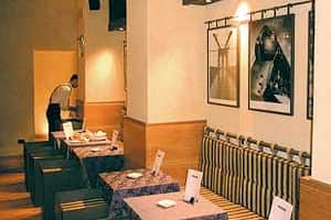 Sala da ristorante con tavoli quadrati e sedie in legno scuro, due quadri in bianco e nero sulla parete.
