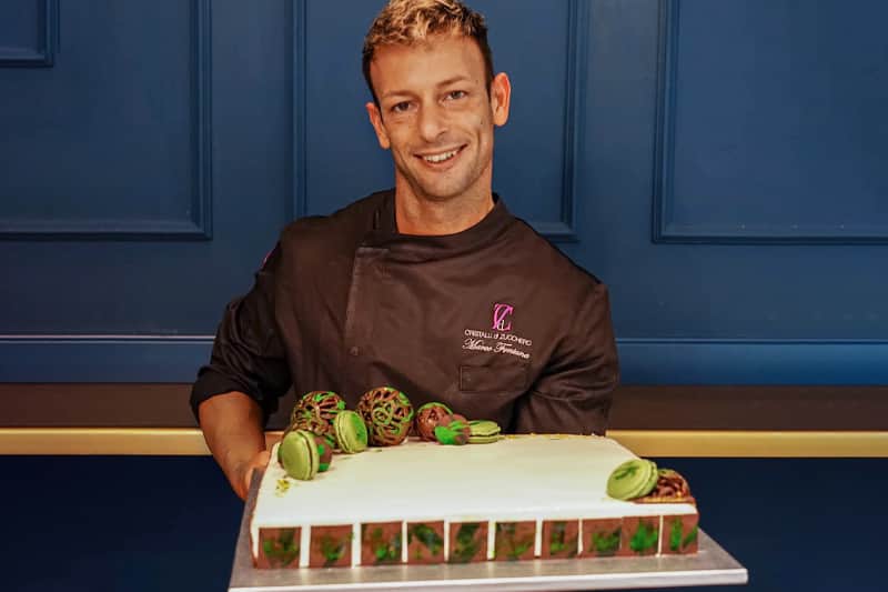 Uomo in giacca da chef sorride reggendo una torta rettangolare con glassa bianca, macaron verdi e sfere di cioccolato.